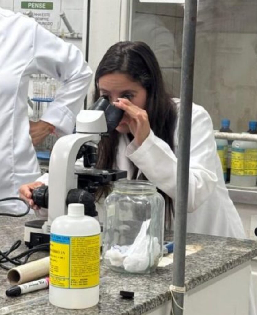 Em um laboratório, uma jovem estudante com cabelos longos e escuros, vestindo jaleco branco, observa atentamente através de um microscópio óptico. Ela está posicionada atrás de uma bancada de pedra, onde também há um pote de vidro contendo papel amassado, uma garrafa plástica com rótulo amarelo, um marcador preto e outros materiais de laboratório. Ao fundo, é possível ver prateleiras com frascos de reagentes e equipamentos científicos. Outra pessoa, também com jaleco branco, está parcialmente visível ao lado esquerdo da imagem. O ambiente é típico de um laboratório de ciências ou biologia, com foco na prática investigativa.