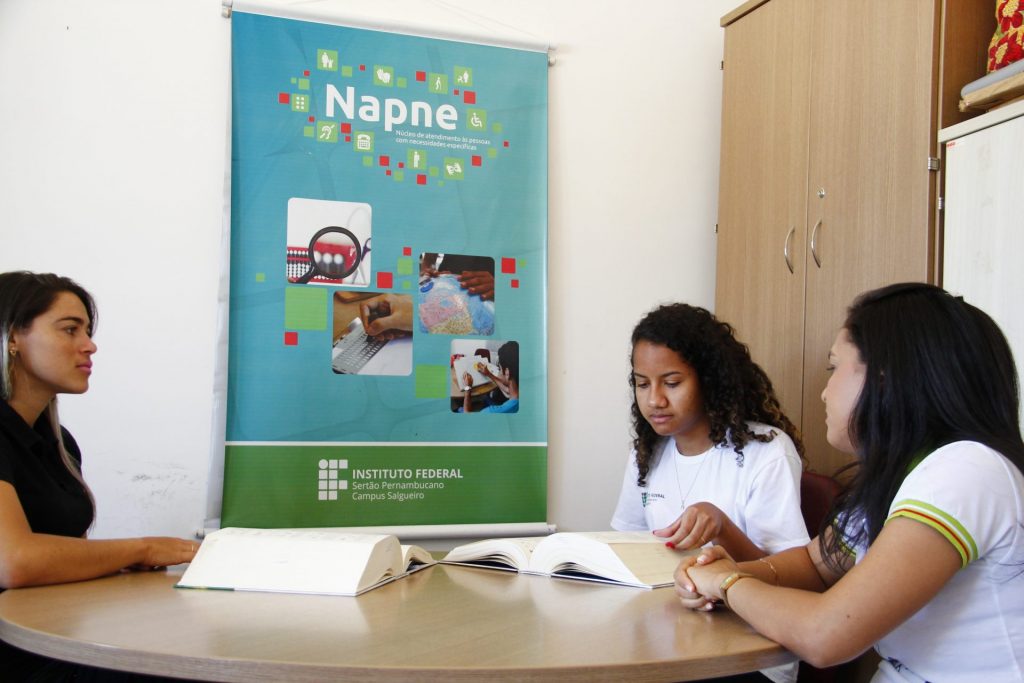 A imagem mostra três mulheres sentadas ao redor de uma mesa redonda em um ambiente interno. Elas estão concentradas, lendo e conversando sobre materiais escritos abertos sobre a mesa. Duas delas usam camisetas brancas com a logomarca do Instituto Federal, indicando que são estudantes. Ao fundo, há um banner vertical do NAPNE — Núcleo de Atendimento às Pessoas com Necessidades Educacionais Específicas — do Instituto Federal do Sertão Pernambucano, Campus Salgueiro. O banner é azul com elementos gráficos coloridos e pequenas imagens que representam inclusão e acessibilidade, como mãos em braille, teclado adaptado, libras e materiais táteis. Ao lado direito da imagem, há um armário de madeira. A cena retrata um momento de atendimento ou orientação pedagógica voltada à inclusão educacional.